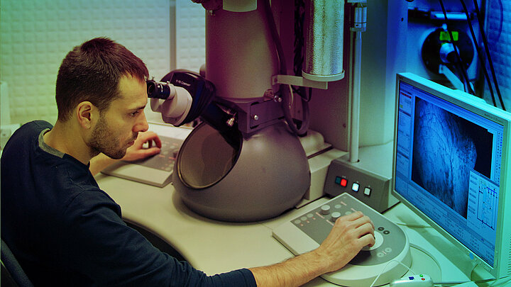 Mężczyzna pracujący przy mikroskopie elektronowym. Ręka spoczywa na pulpicie.