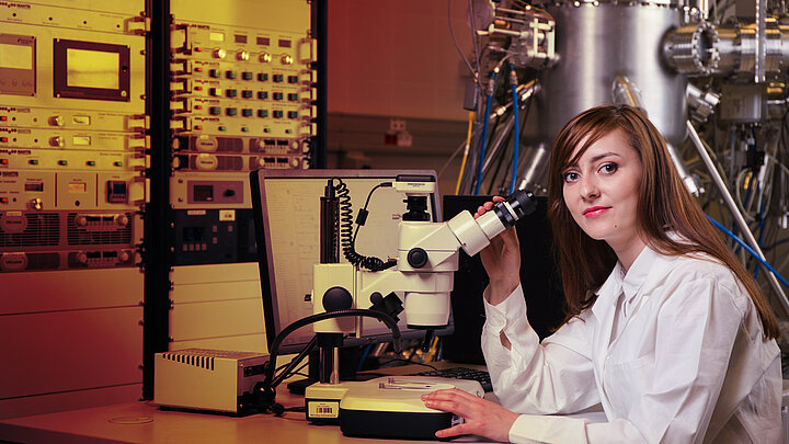 Młoda kobieta naukowiec siedzi przy biurku z mikroskopem. W tle skomplikowana aparatura badawcza.