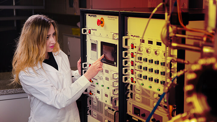 Młoda naukowczyni stoi obok szaf z aparaturą badawczą. Palcem wskazuje na dane przezentowane na wyświetlaczu.